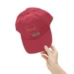 Red baseball cap being held by a hand against a white background, viewed from the side.