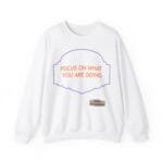 White sweatshirt featuring an orange slogan 'FOCUS ON WHAT YOU ARE DOING' inside a purple decorative frame, with a small beige label reading 'Design By The Simple MindSet' near the hem.