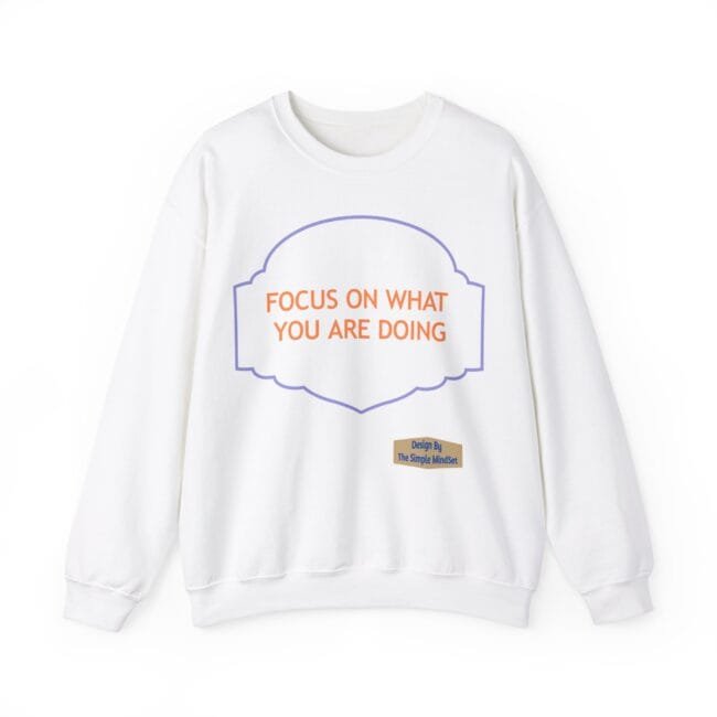 White sweatshirt featuring an orange slogan 'FOCUS ON WHAT YOU ARE DOING' inside a purple decorative frame, with a small beige label reading 'Design By The Simple MindSet' near the hem.