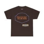 Brown T-shirt with orange text 'FOCUS ON WHAT YOU ARE DOING' inside a purple decorative frame; small beige designer label reading 'Designed By The Simple Design MindSet'