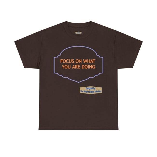 Brown T-shirt with orange text 'FOCUS ON WHAT YOU ARE DOING' inside a purple decorative frame; small beige designer label reading 'Designed By The Simple Design MindSet'
