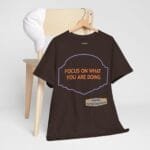 Brown T-shirt on a hanger with a purple framed badge reading 'FOCUS ON WHAT YOU ARE DOING' in orange letters.
