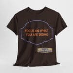 Dark brown T-shirt featuring an orange message 'FOCUS ON WHAT YOU ARE DOING' inside a purple decorative frame; a beige label reads 'Designed By The Simple Design MindSet' at the bottom right.