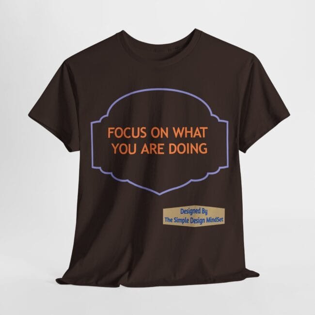 Dark brown T-shirt featuring an orange message 'FOCUS ON WHAT YOU ARE DOING' inside a purple decorative frame; a beige label reads 'Designed By The Simple Design MindSet' at the bottom right.
