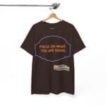 Brown T-shirt on a wooden hanger with orange text 'FOCUS ON WHAT YOU ARE DOING' inside a purple decorative frame; small beige designer label reads 'Designed By The Simple Design Mindset' at the bottom.