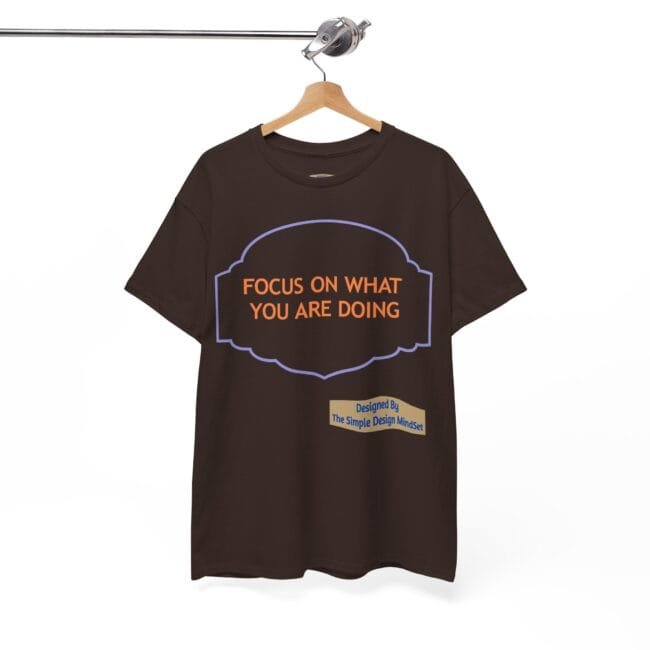 Brown T-shirt on a wooden hanger with orange text 'FOCUS ON WHAT YOU ARE DOING' inside a purple decorative frame; small beige designer label reads 'Designed By The Simple Design Mindset' at the bottom.