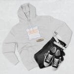 Flat lay of a light gray hoodie with a decorative label on the chest, paired with dark jeans and black‑and‑white sneakers on a light background.