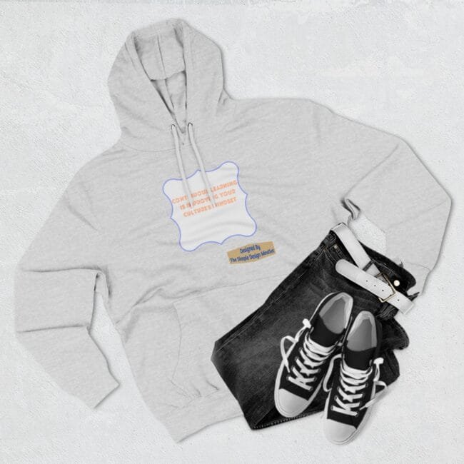 Flat lay of a light gray hoodie with a decorative label on the chest, paired with dark jeans and black‑and‑white sneakers on a light background.