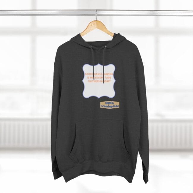 Dark gray hoodie on a hanger with a pale blue decorative label containing orange text about continuous learning and culture's mindset; small beige designer label at bottom right.