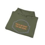 Olive green folded hoodie featuring a lavender cloud border and orange text: 'FOCUS ON WHAT YOU ARE DOING'.