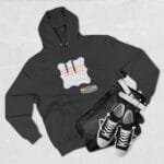 Flat lay of a dark hoodie with a white decorative label and orange text, paired with blue jeans, a white belt, and black-and-white sneakers nearby.