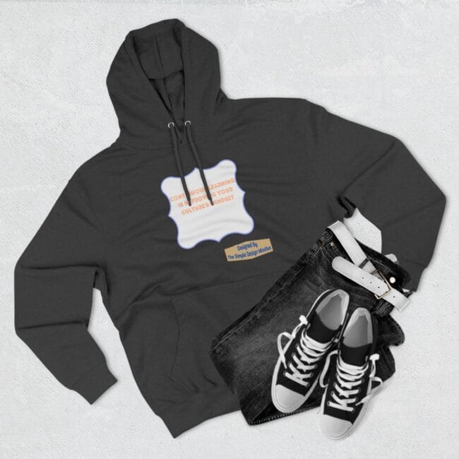Flat lay of a dark hoodie with a white decorative label and orange text, paired with blue jeans, a white belt, and black-and-white sneakers nearby.