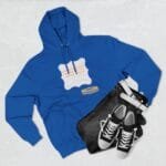 Flat lay of a royal blue hoodie with a white decorative patch and orange text, paired with black jeans and white‑trim sneakers on a light background.