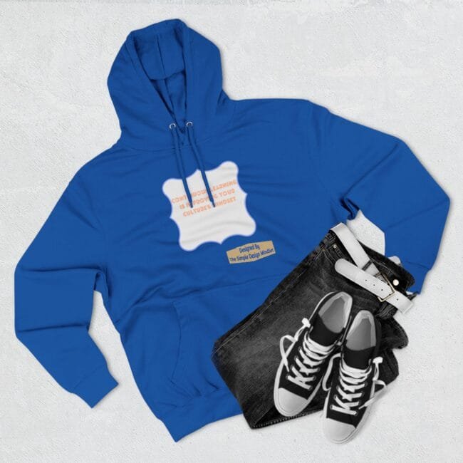 Flat lay of a royal blue hoodie with a white decorative patch and orange text, paired with black jeans and white‑trim sneakers on a light background.