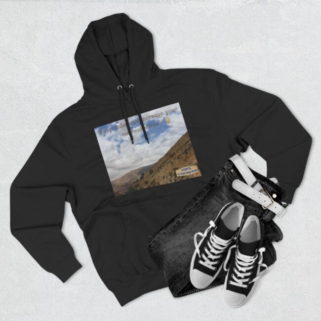 Black hoodie laid flat with a rectangular mountain landscape graphic on the chest and faint inspirational text across the top of the image area, paired with jeans and sneakers as accessories in the scene.