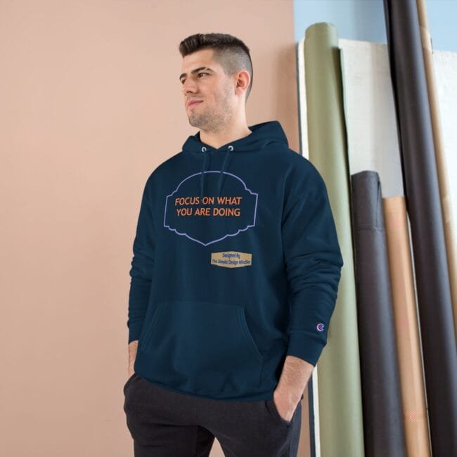 Man wearing a navy hoodie with orange text 'FOCUS ON WHAT YOU ARE DOING' standing beside rolled fabrics in a studio setting, hands in pockets.