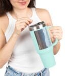 Person holding a light blue insulated tumbler with a straw, preparing to sip from it while wearing a white tank top and jeans.