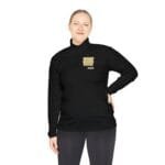 Woman in a black long-sleeve pullover with a zipped collar, standing with one hand on her hip and a small badge on the left chest.