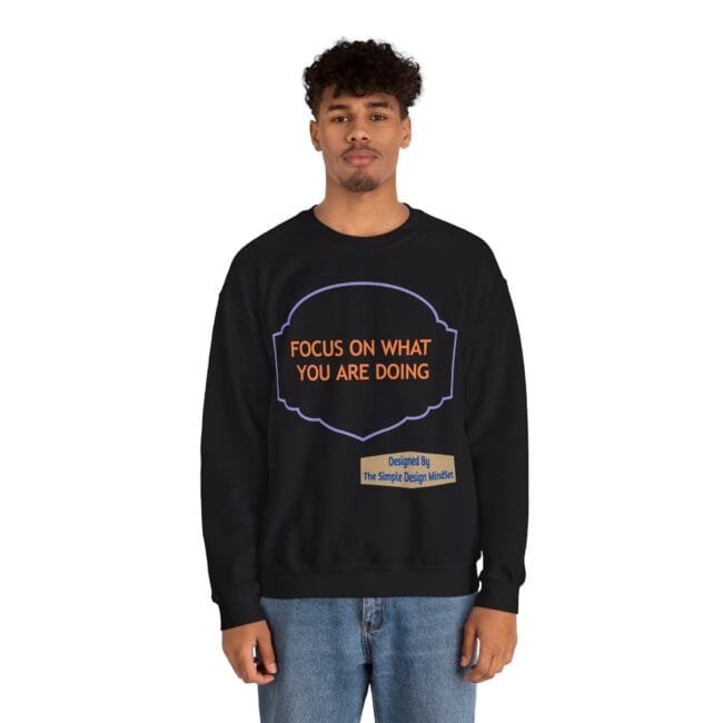 Young man wearing a black sweatshirt with orange text 'FOCUS ON WHAT YOU ARE DOING' inside a purple badge, plus a small beige label reading 'Designed By The Simple Design Mindset'