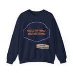 Navy sweatshirt with orange motto 'FOCUS ON WHAT YOU ARE DOING' in a purple decorative frame and a beige credit banner below.