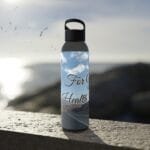 Metal water bottle with a black cap sits on a sunlit concrete ledge; label shows a blue sky with clouds and a road, with the words 'For Health' visible.