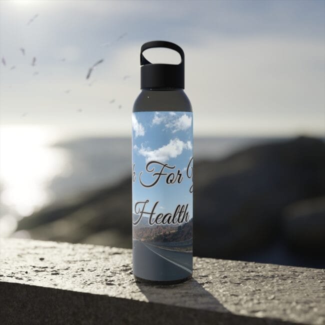 Metal water bottle with a black cap sits on a sunlit concrete ledge; label shows a blue sky with clouds and a road, with the words 'For Health' visible.