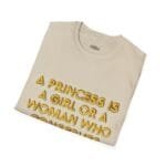 Beige folded T-shirt with gold 3D lettering that reads: “A princess is a girl or a woman who ...”