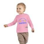 Cute toddler in a pink long-sleeve shirt that reads 'Take care of my PARENTS' in blue, smiling and walking with a small object in hand.
