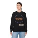 Woman wearing a black sweatshirt with an orange slogan: ‘FOCUS ON WHAT YOU ARE DOING’ inside a purple border.