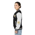 Side view of a young woman wearing a black and gray varsity jacket with white striped cuffs and patches on the chest and sleeve.
