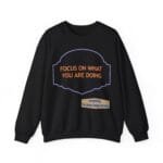 Black sweatshirt with an orange quote 'FOCUS ON WHAT YOU ARE DOING' inside a purple decorative frame and a beige designer tag at the bottom left.