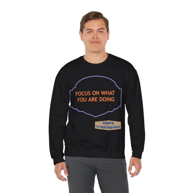 Man wearing a black sweatshirt with an orange slogan: 'FOCUS ON WHAT YOU ARE DOING' inside a purple outlined badge; small designed-by tag bottom right