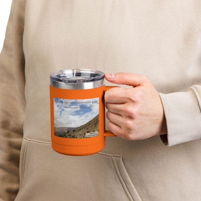 Close-up of a hand gripping an orange insulated mug with a clear lid and a landscape sticker reading 'Keep calm and increase your concentration'.