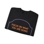 Black folded T-shirt with orange slogan 'FOCUS ON WHAT YOU ARE DOING' inside a purple decorative border.