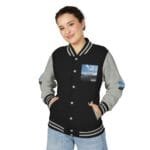 Young woman in a black and gray varsity jacket with patches, smiling at the camera against a white studio background, hands in pockets.