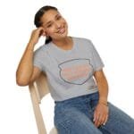 Smiling woman seated on a wooden chair, wearing a gray T-shirt with an orange slogan: 'Keep calm and don't disturb my head please!'.
