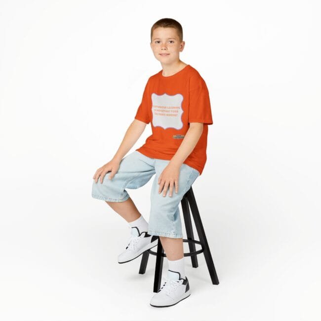 Young boy sits on a black stool in a studio, wearing an orange T-shirt, light denim shorts, white socks and sneakers against a white background.