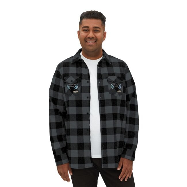 Man wearing a black and gray plaid flannel shirt over a white tee, smiling at the camera with embroidered patches on each chest pocket.