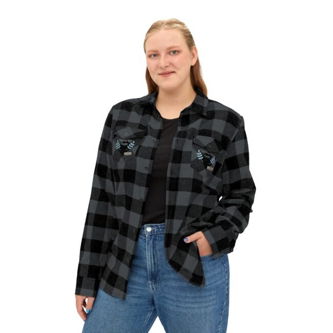 Woman wearing a black-and-gray plaid flannel shirt with embroidered patches on the chest pockets, posing with one hand in her pocket.