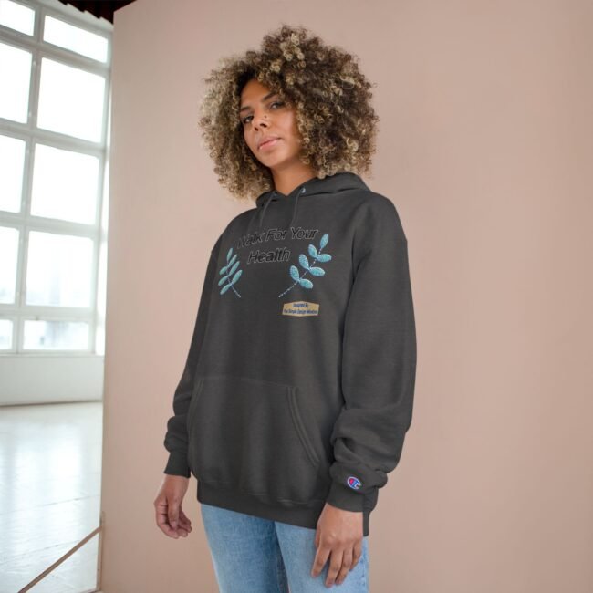 Woman in a dark hoodie with teal leaf graphics and the text 'Walk For Your Health', standing indoors against a pale pink wall by a large window.