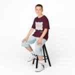 Smiling boy sits on a black stool, wearing a burgundy T-shirt with a light-blue graphic and light-wash jeans against a white background.