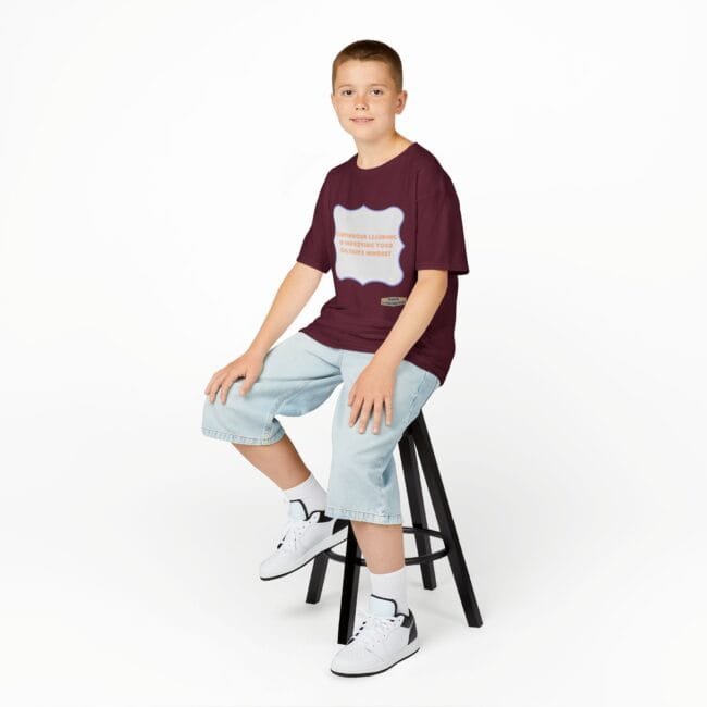 Smiling boy sits on a black stool, wearing a burgundy T-shirt with a light-blue graphic and light-wash jeans against a white background.