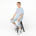 Smiling boy sits on a black stool in a light blue graphic T-shirt, light denim shorts, and white sneakers against a white studio background.