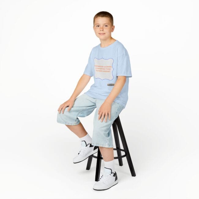 Smiling boy sits on a black stool in a light blue graphic T-shirt, light denim shorts, and white sneakers against a white studio background.