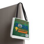Gray reusable tote bag with black straps hanging from a corner, showing a green poster that reads 'World Cup 2026' with soccer imagery and a trophy
