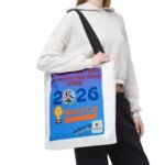 Person in a white hoodie carries a bright blue poster with a soccer ball and the year 2026, strapped over the shoulder with a black bag strap