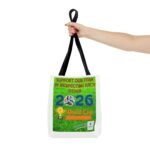 Hand gripping the black straps of a white tote bag with a green poster: 'Support our team by respecting each other' and soccer imagery on grass background